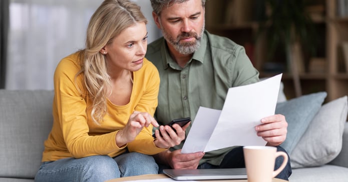 A couple sitting on their couch reviewing bills and calculating expenses, reflecting on homeownership affordability.